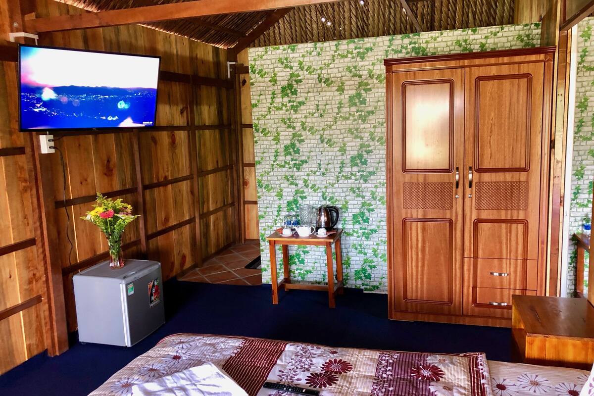 Habitació amb parets de fusta, televisió penjada a la paret, llit amb colcha floral, taula petita amb tetera i tasses, i armari de fusta.
