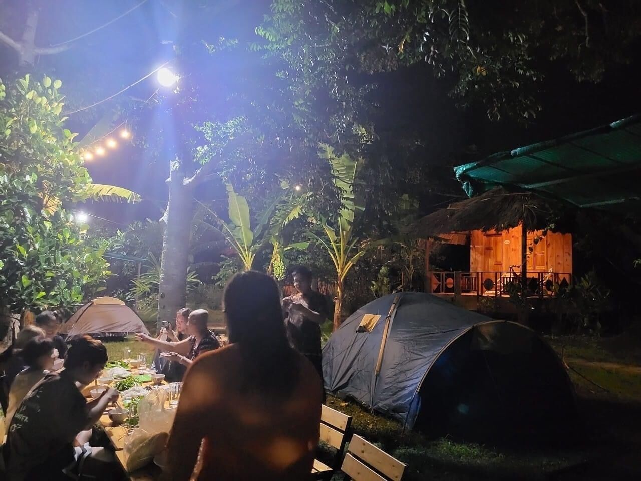 Eine Gruppe von Menschen speist draußen bei Nacht, umgeben von Zelten und einer Holzhütte mit einem Strohdach.