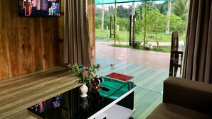 Una sala de estar moderna con un televisor montado en la pared, una mesa de centro de vidrio y vistas a un jardín a través de grandes ventanales.
