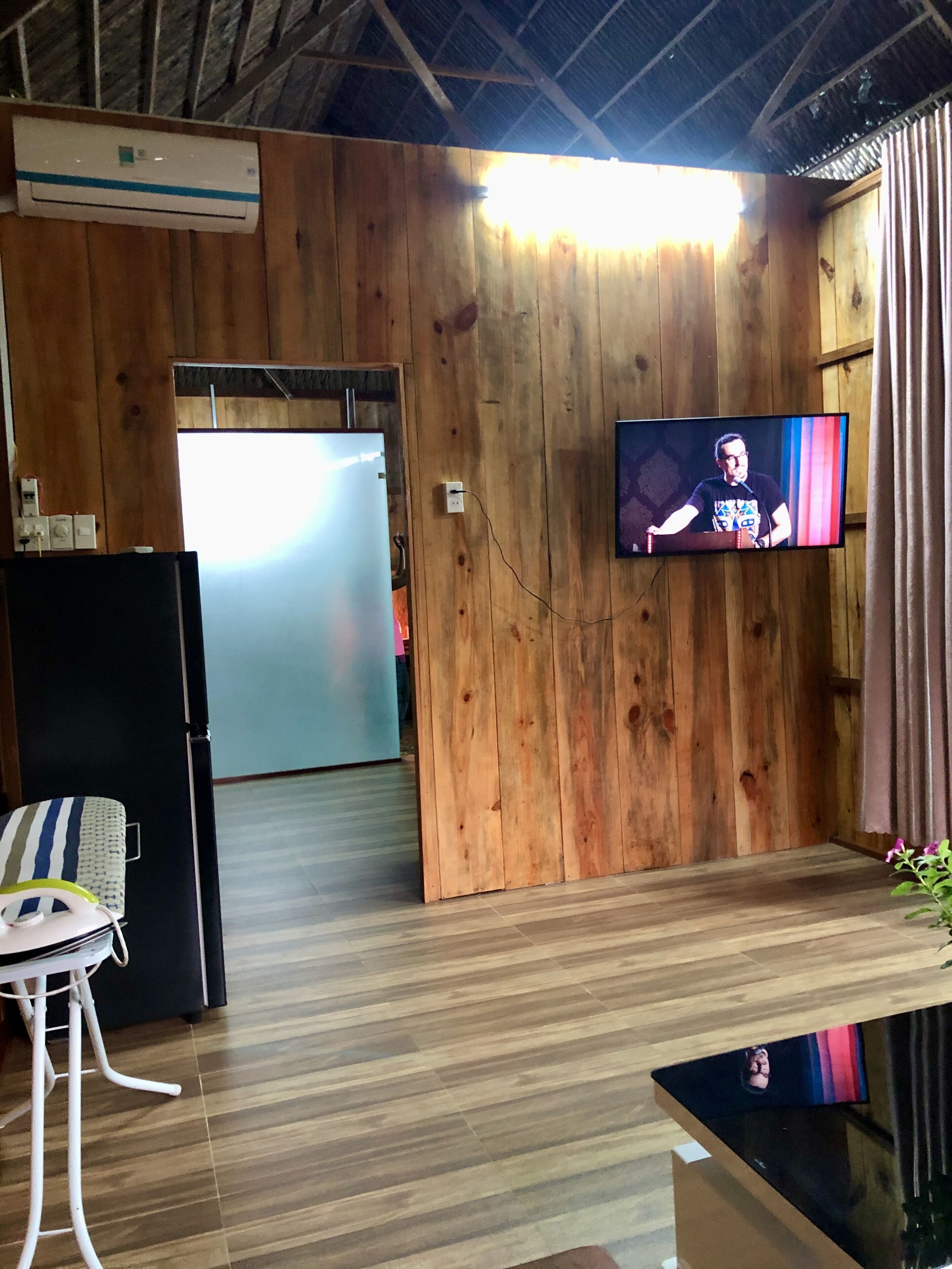 Quarto com paredes de madeira, uma televisão de tela plana presa à parede, um refrigerador e uma cadeira com almofada listrada.