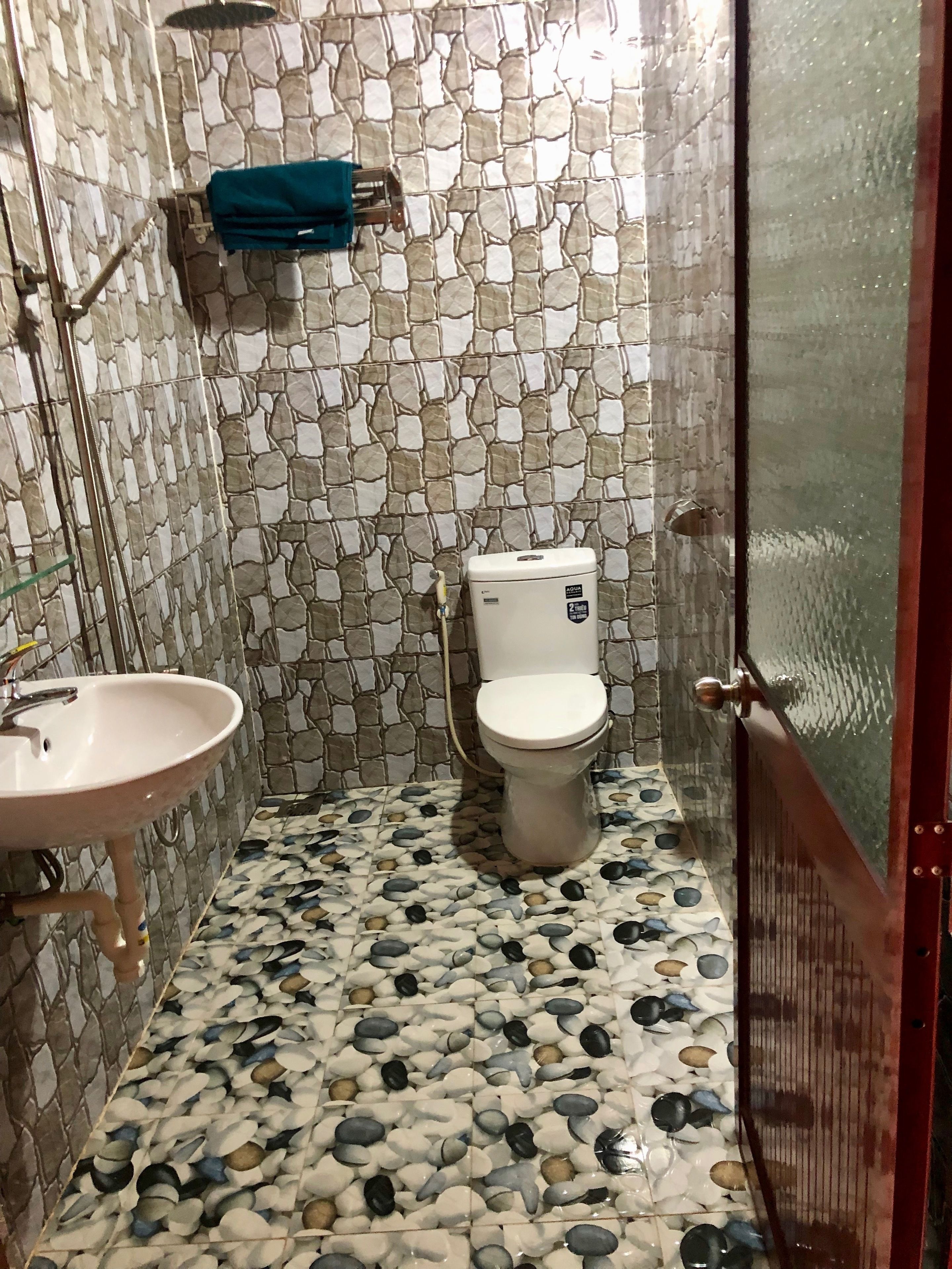A bathroom with a stone mosaic wall, a white toilet, and a sink with a blue towel hanging on the wall.