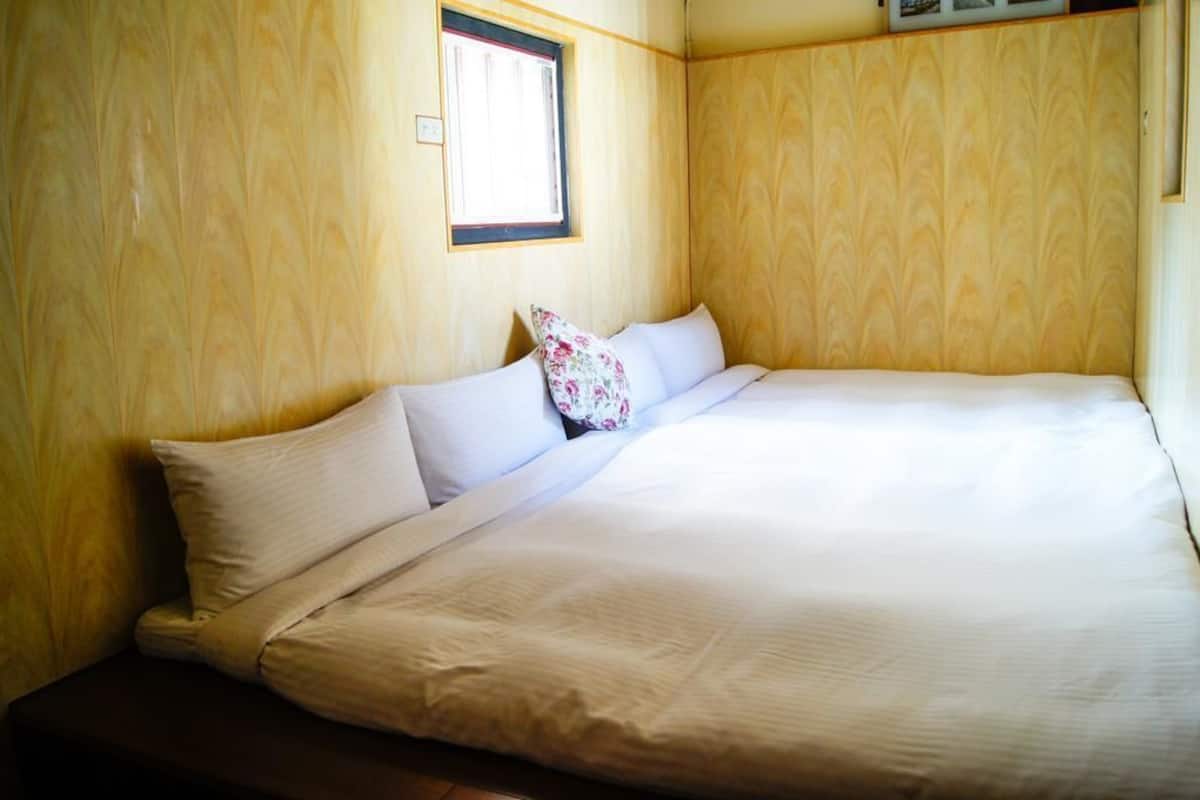 A single bed with white linens and a floral pillow, set against a wooden wall with a window above it.