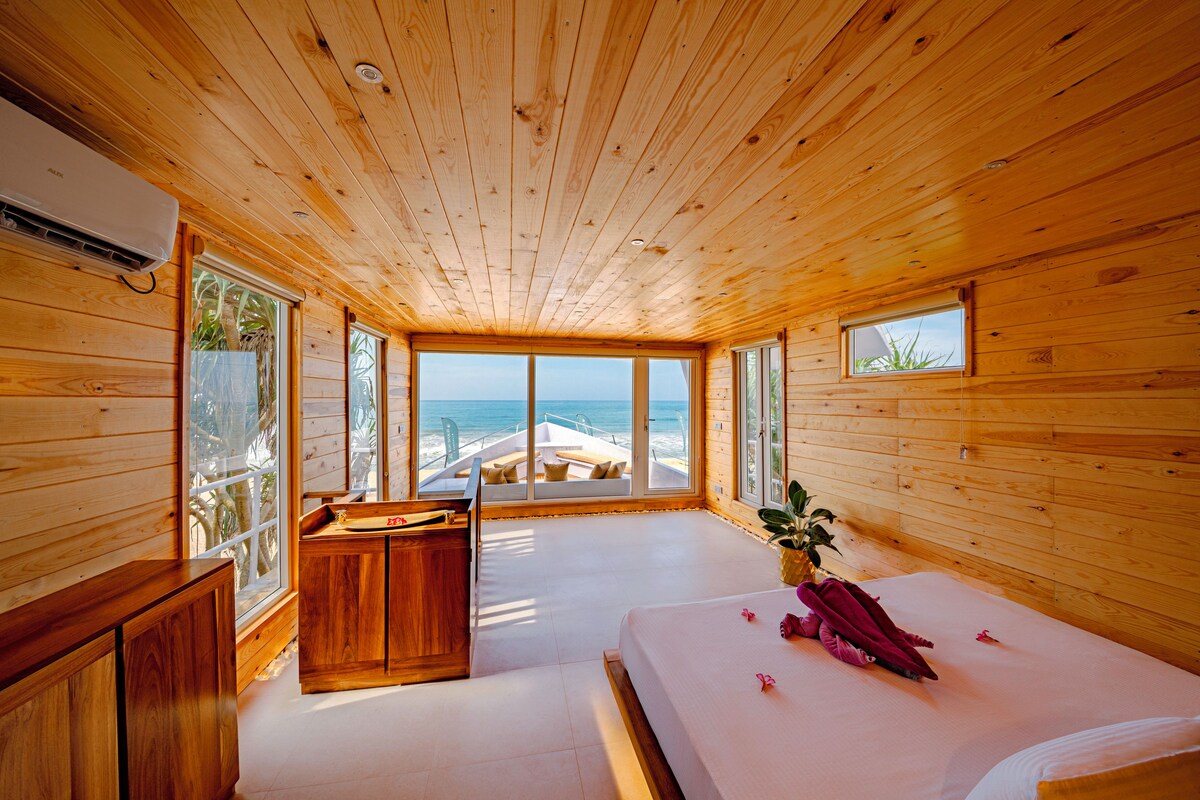 A wooden cabin with a large bed, a wooden cabinet, and a view of the beach.