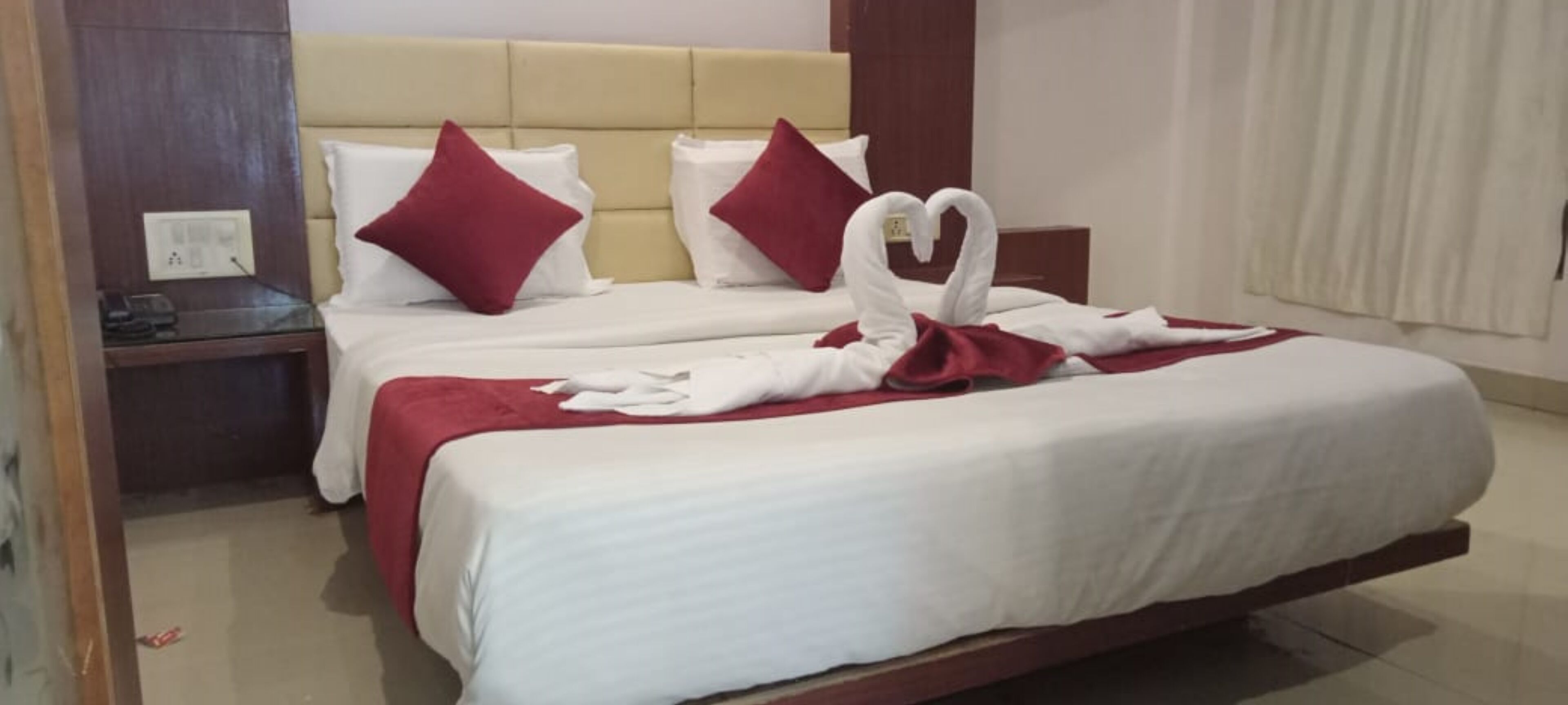 A hotel room with a bed featuring white bedding, red pillows, and a red blanket. A towel is arranged in a heart shape on the bed.