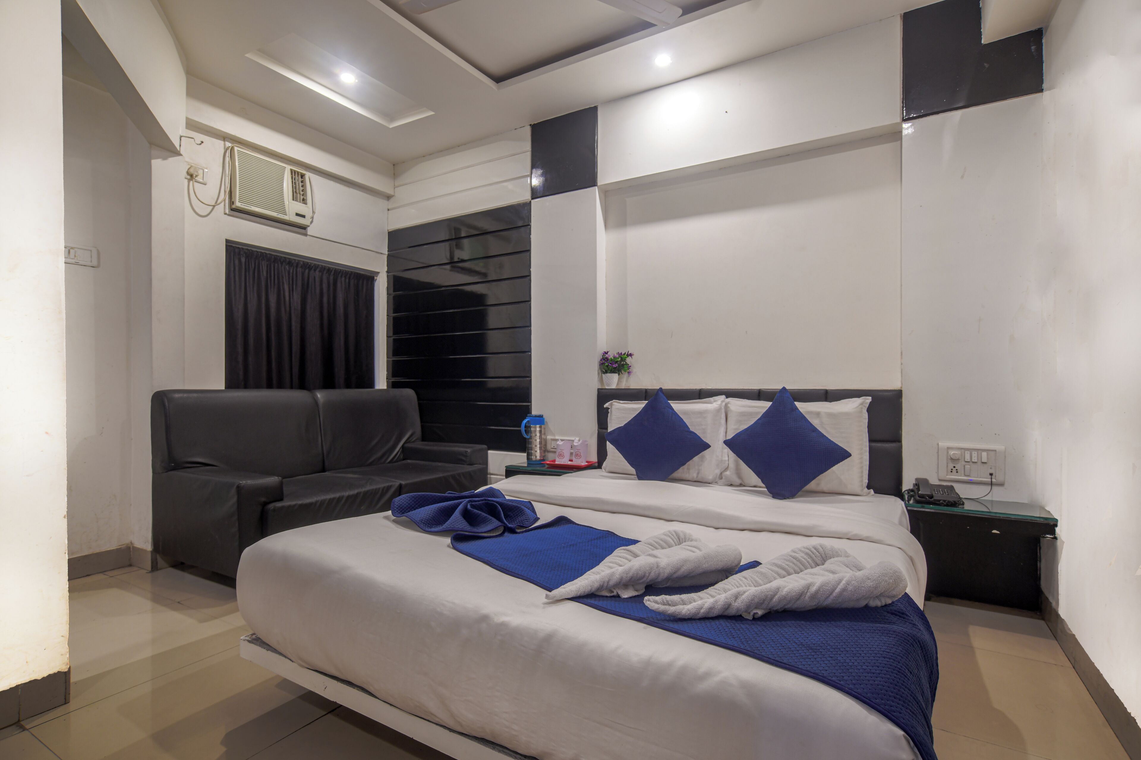 A hotel room with a large bed, a sofa, a small table, and a wall-mounted air conditioning unit.