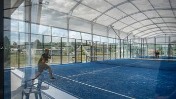 A tennis court with a net, surrounded by glass walls and a metal roof, offering views of the surrounding area.
