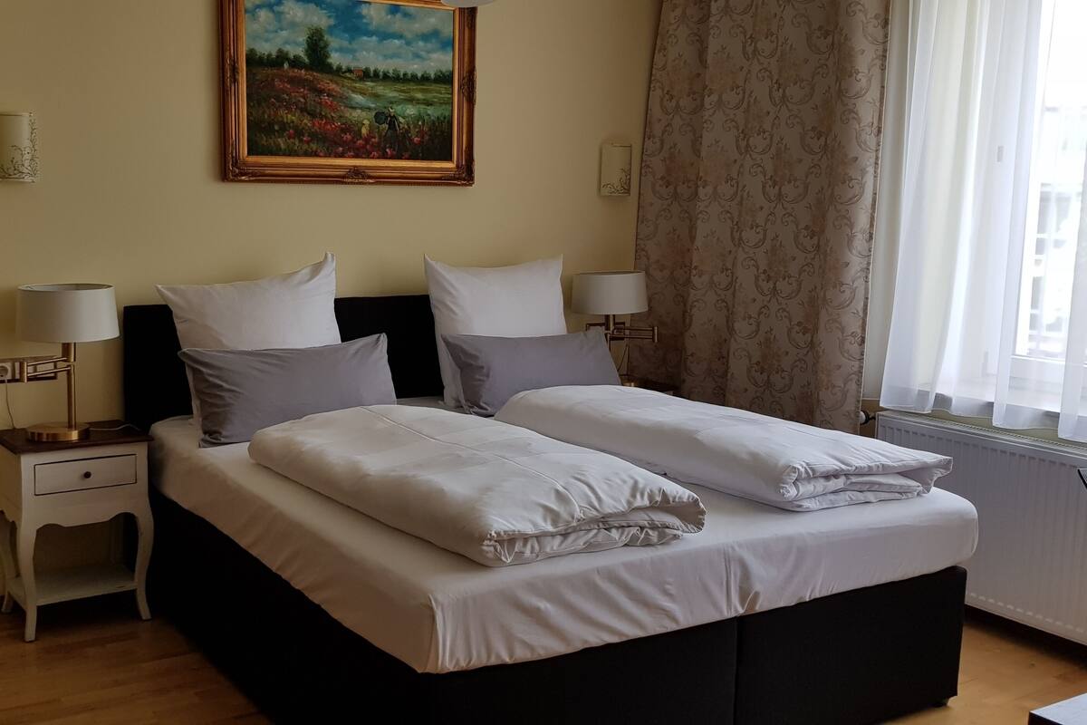 Un letto matrimoniale con lenzuola bianche, due comodini con lampade, un quadro appeso alla parete e una finestra con tende.