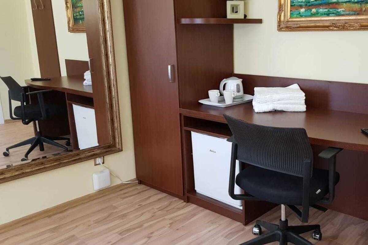 Una camera d'albergo con una scrivania in legno, una sedia da ufficio nera, un piccolo frigorifero, uno specchio, un quadro appeso al muro e un cestino nero.
