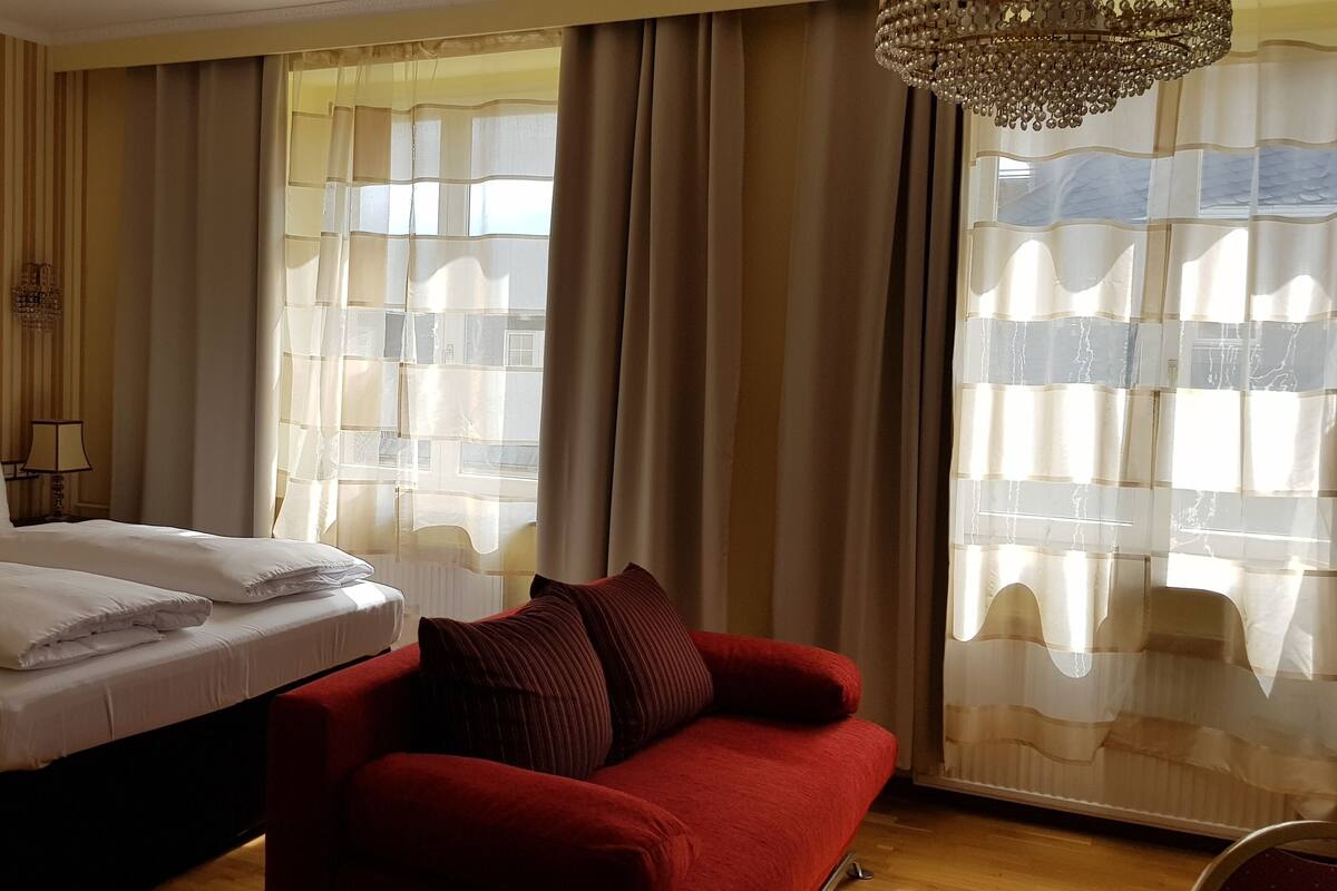 Una camera d'albergo con un letto, un divano rosso, un lampadario e ampie finestre con tende.
