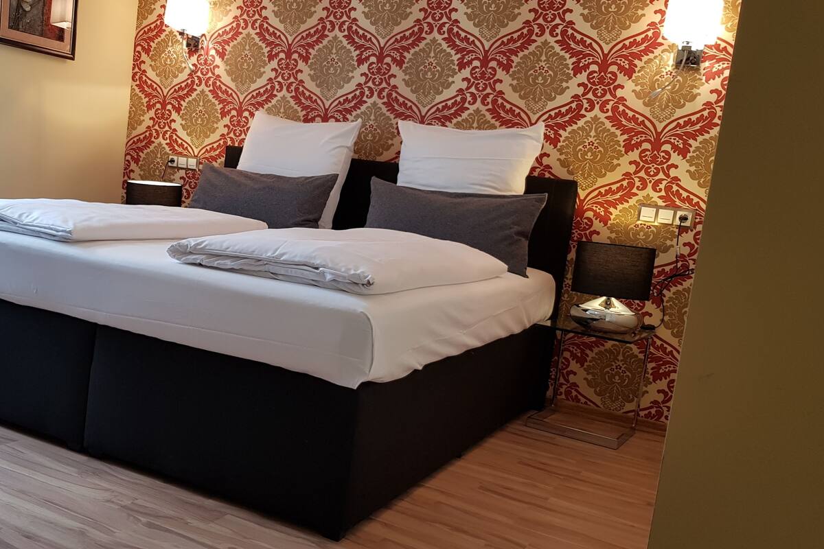 Un letto rifatto con lenzuola e cuscini bianchi, una testata scura e una parete con carta da parati decorata.