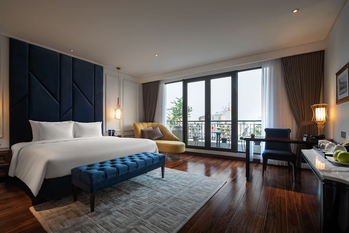 A modern hotel room with a large bed, a sitting area with a chair and a desk, a balcony with a view, and a large window.