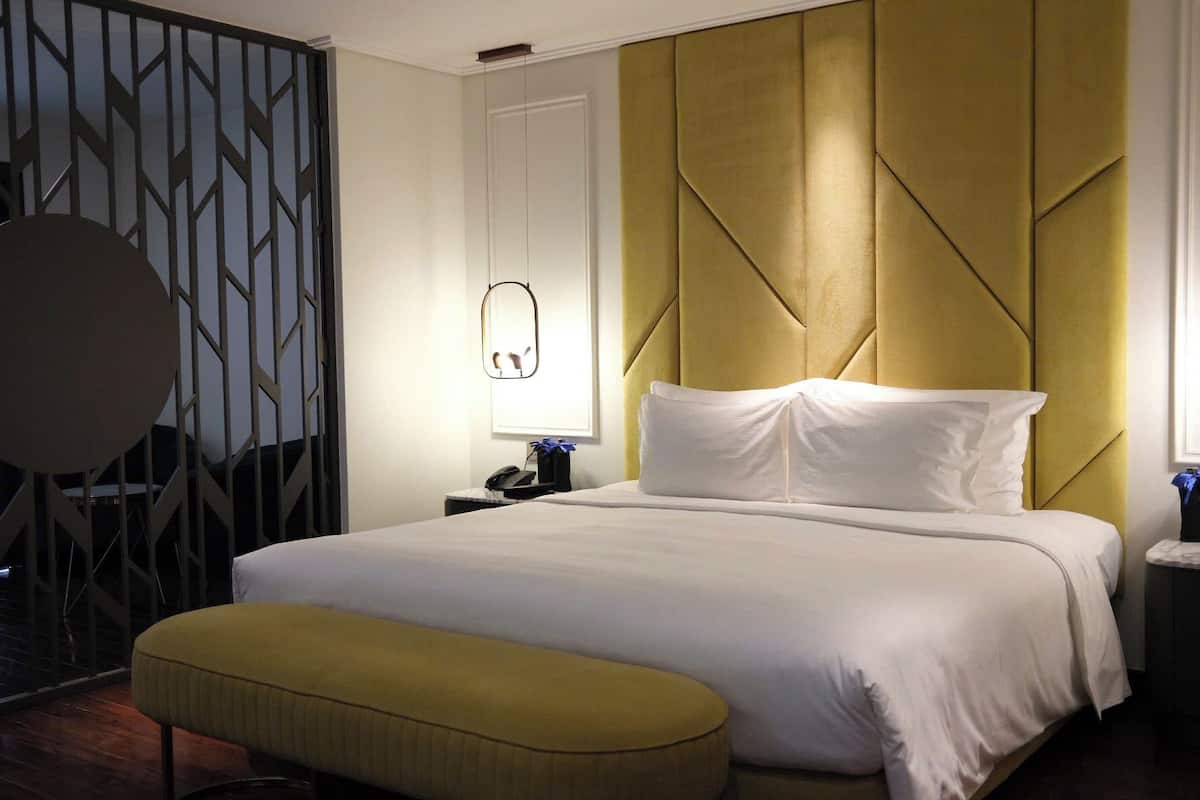 A hotel room with a large bed, a bedside table, a nightstand, a wall-mounted mirror, and a decorative partition.