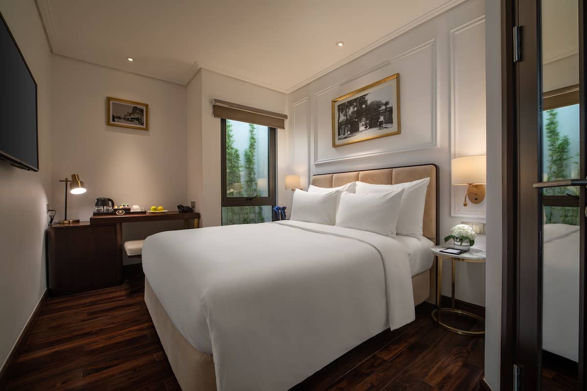 A hotel room with a large bed, a desk with a lamp, a small table with a flower, and a window with a view of trees.