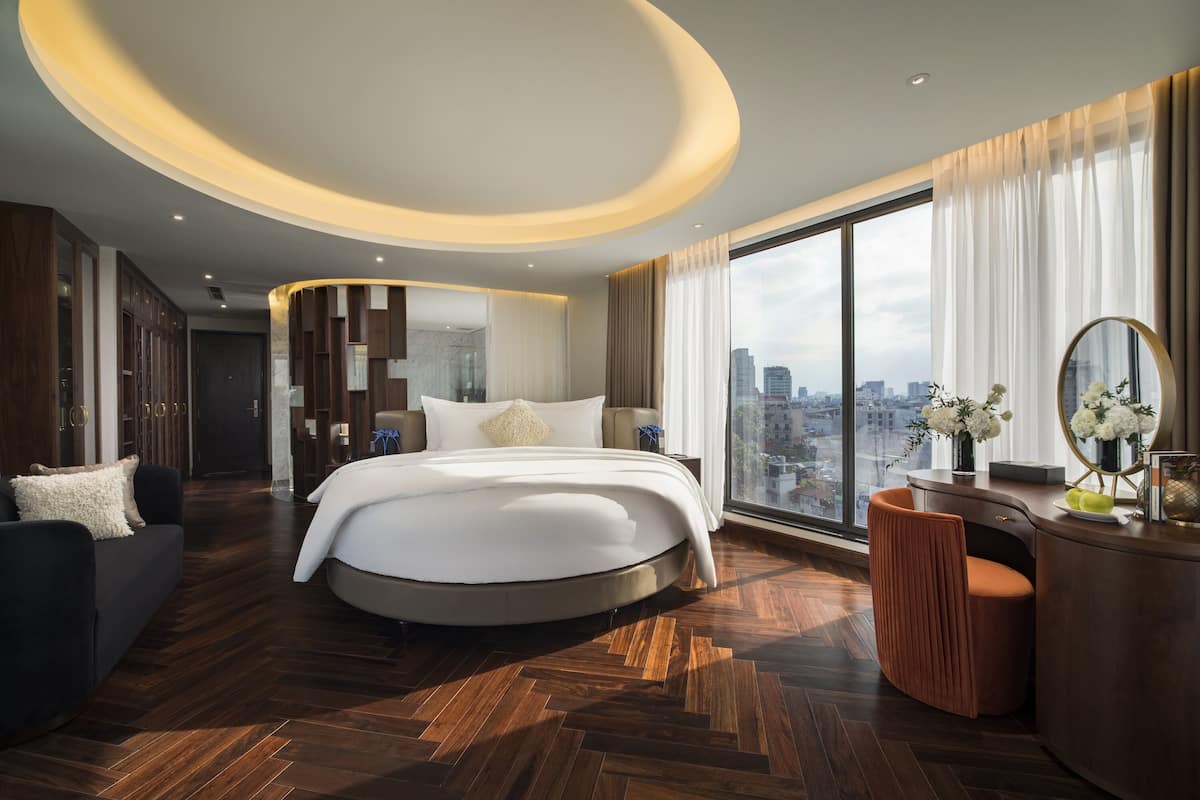 A modern hotel room with a large bed, a round desk, and a view of the city.