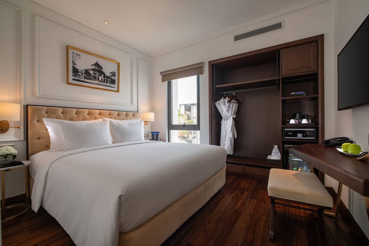 A hotel room with a large bed, a wooden desk, a robe hanging on a rack, and a television on the wall.