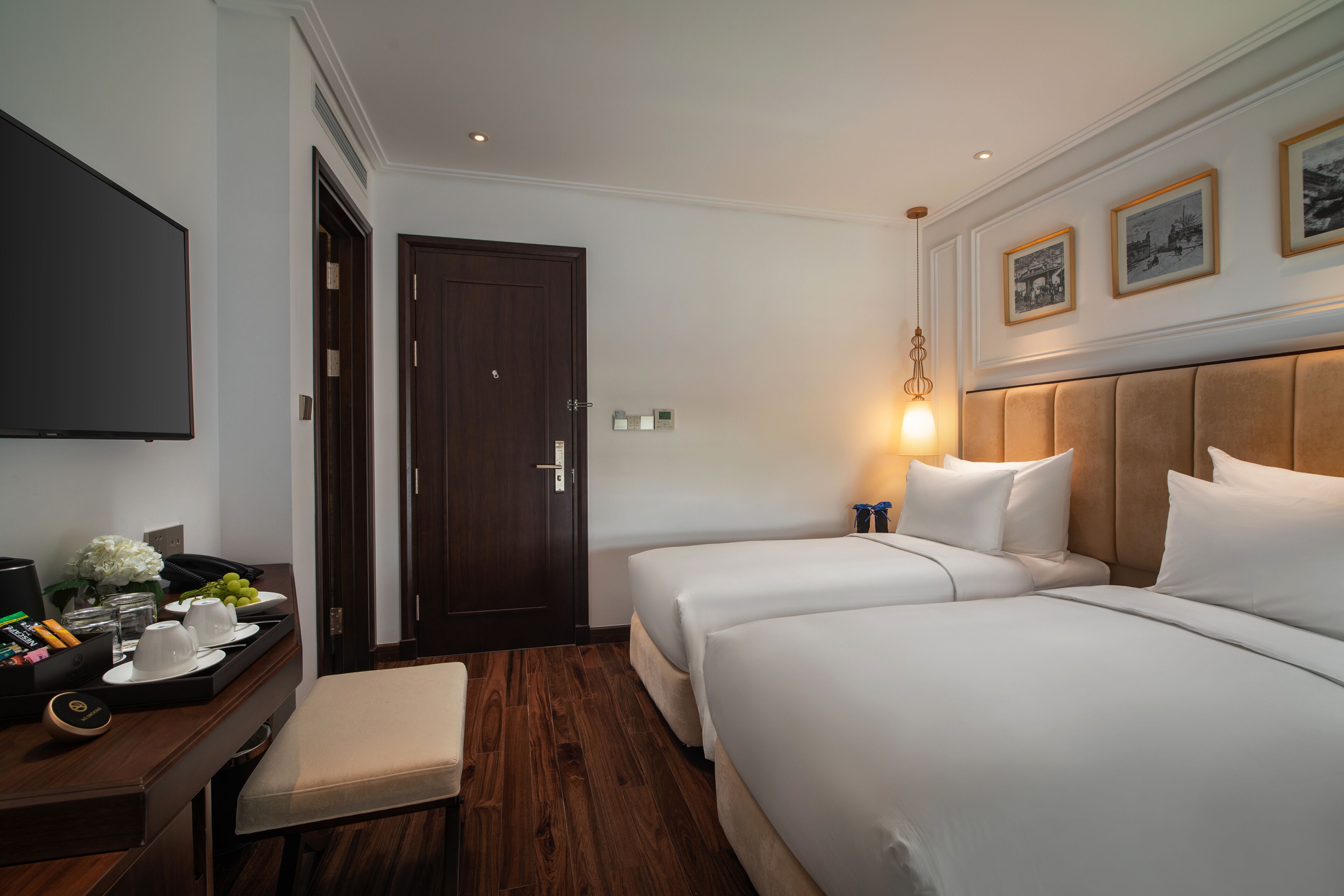 A hotel room with two beds, a desk with a tray of breakfast items, a flat-screen TV, and a wooden floor.