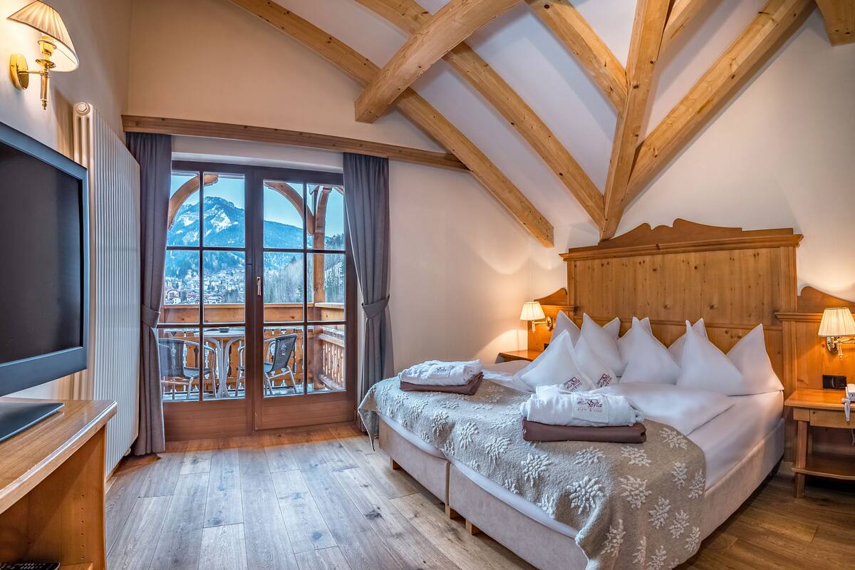 Une chambre avec un grand lit, des poutres apparentes, une télévision et un balcon offrant une vue sur les montagnes.