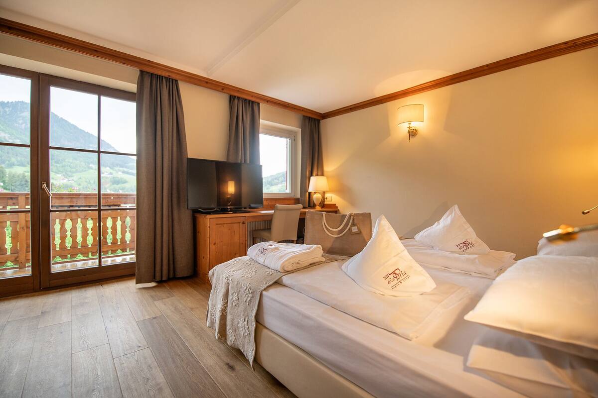 Une chambre d’hôtel avec un balcon, une télévision, un bureau avec une lampe et des lits bien faits, avec des oreillers.
