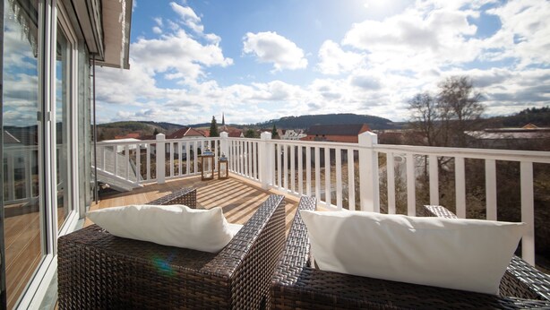 A balcony with wicker furniture and a white railing, overlooking a scenic landscape with hills and a church. - Haus Grünewald Unsleben (Unsleben)
