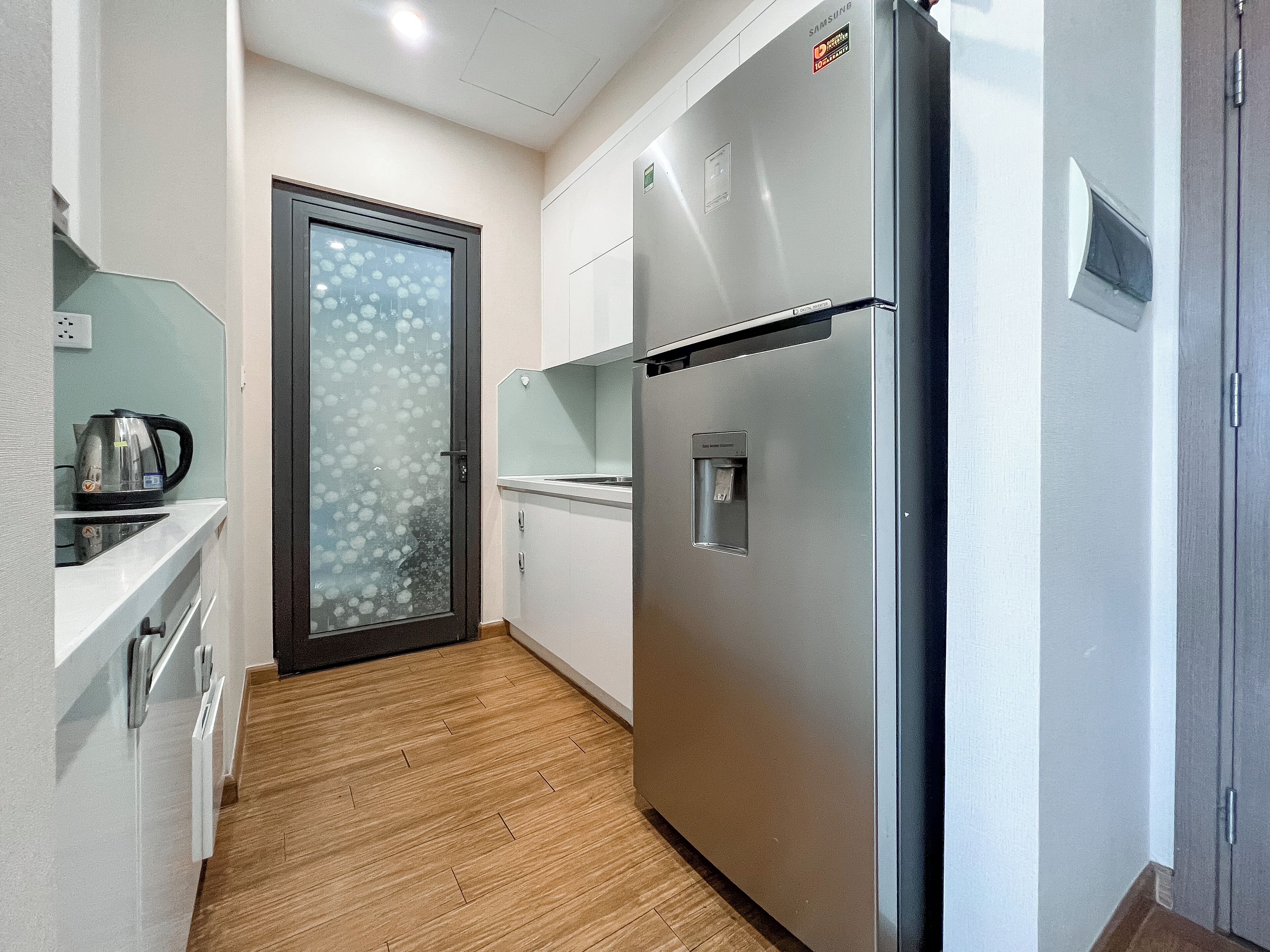 A compact kitchen with a stainless steel refrigerator, a kettle on the counter, and a frosted glass door.