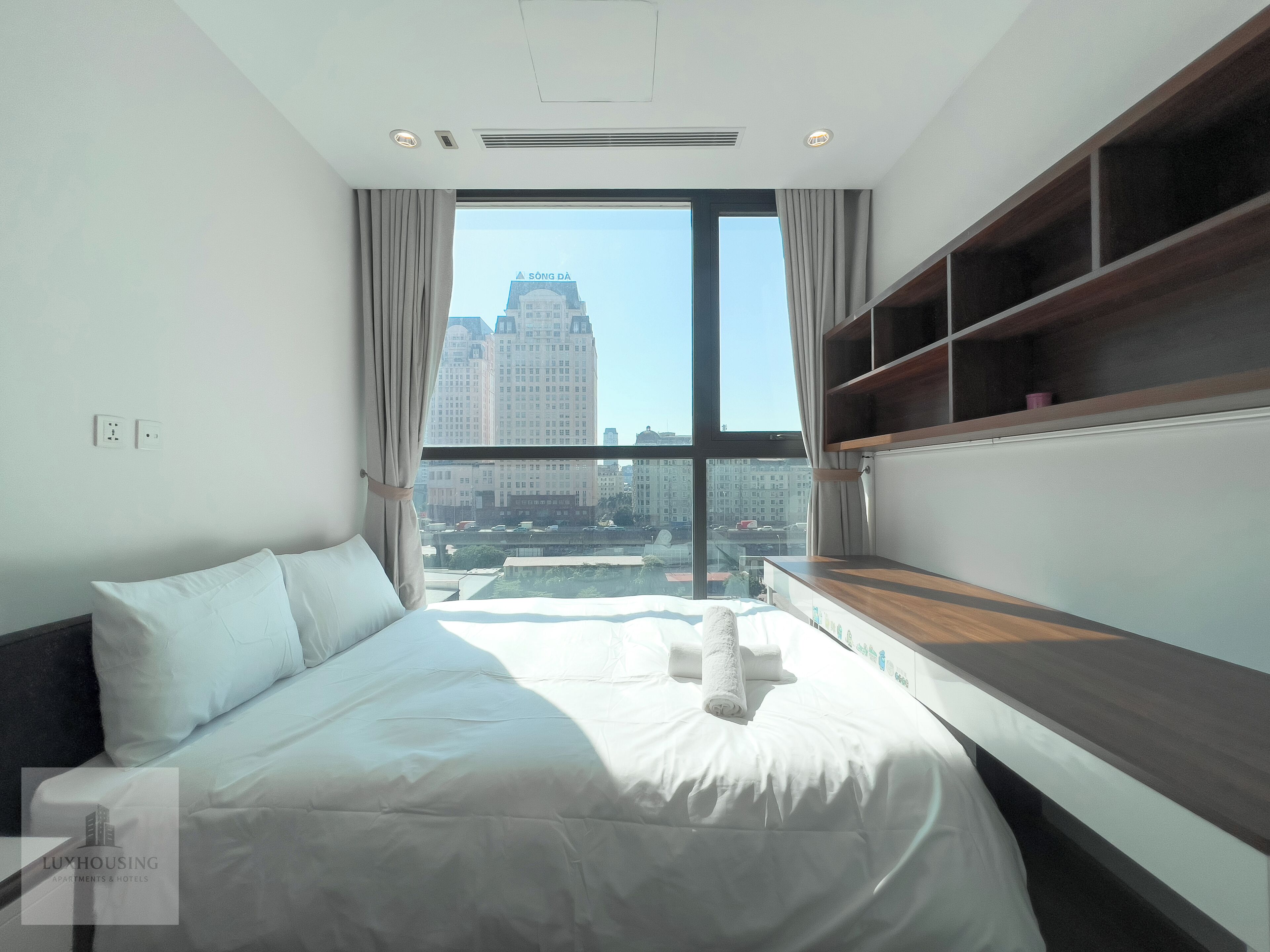 A modern hotel room with a large bed, a built-in shelf, and a city view.