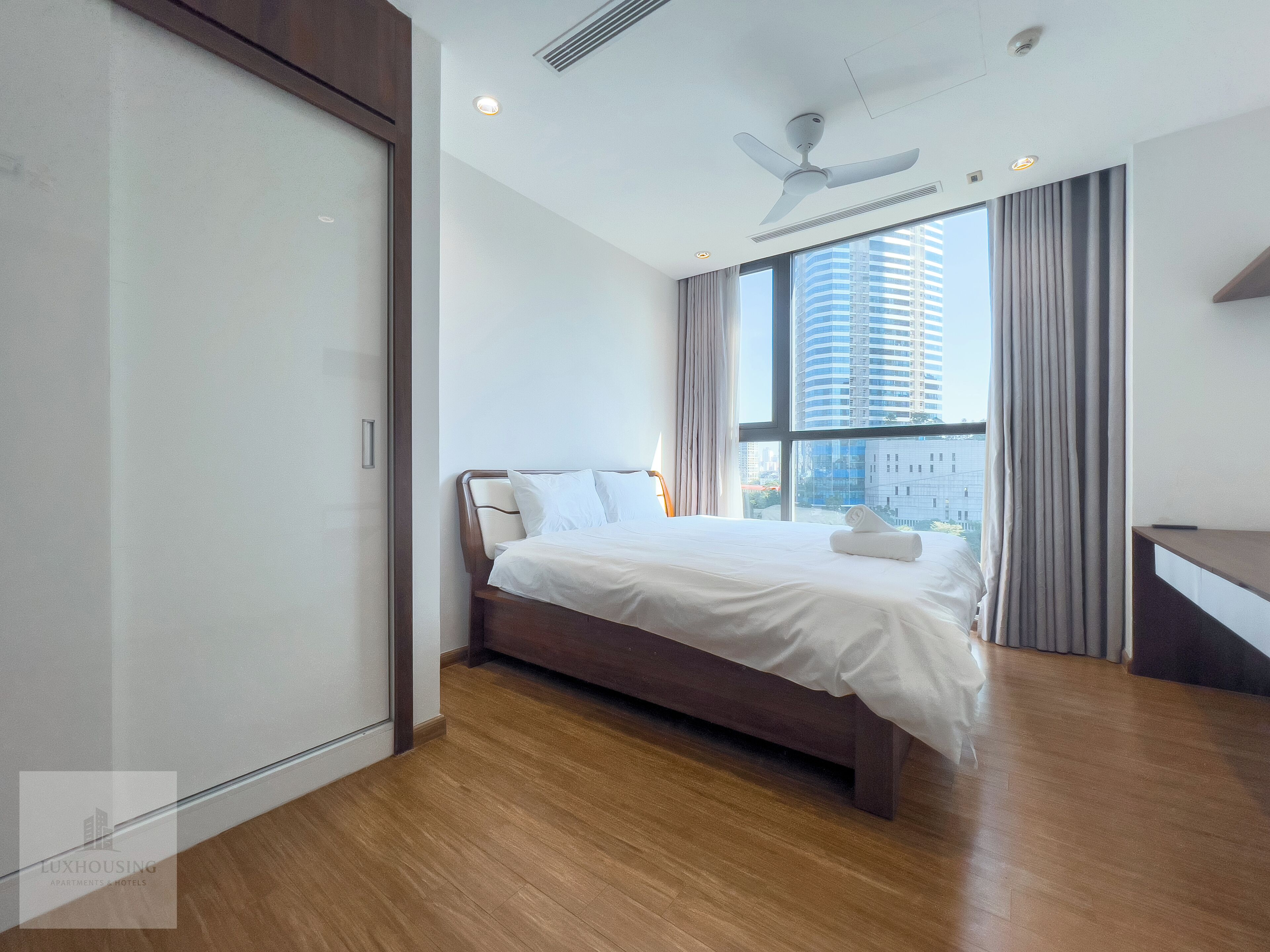 A modern hotel room with a large bed, a ceiling fan, a window with a city view, and a wooden floor.