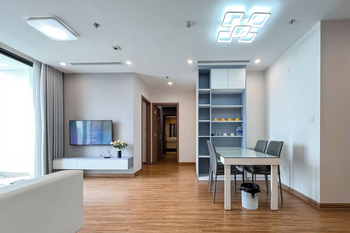 A modern living room with a flat-screen TV, a white sofa, a dining table with chairs, and a kitchen area with shelves.