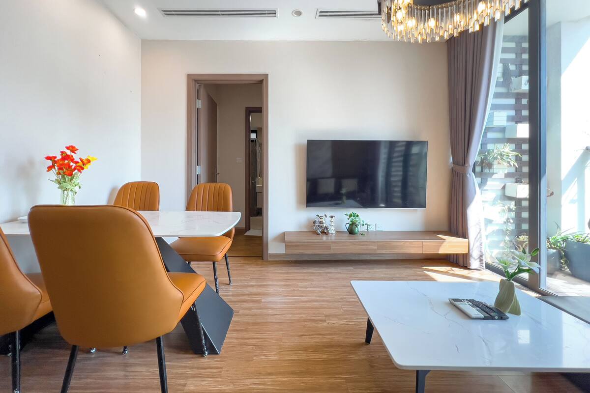 A modern living room with a dining area, a flat-screen TV, and a large window with curtains.