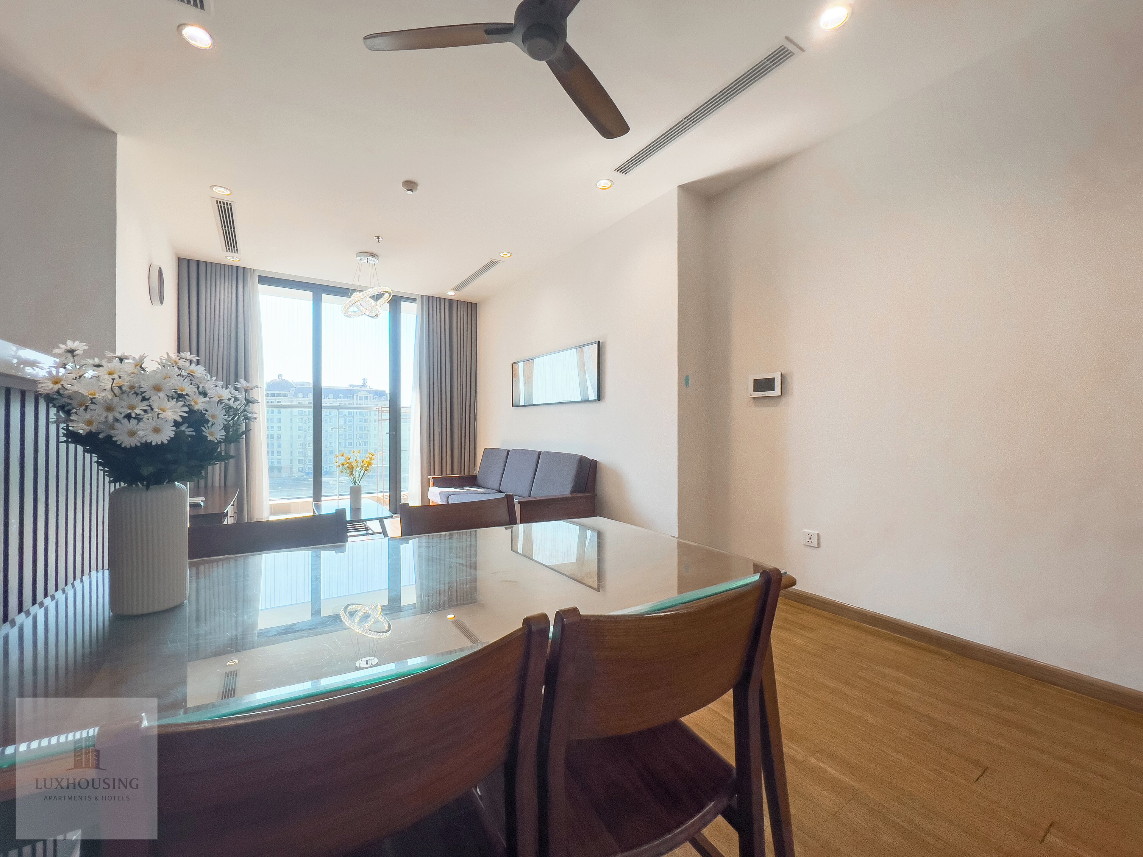 A modern hotel room with a glass dining table, a sofa, and a ceiling fan.