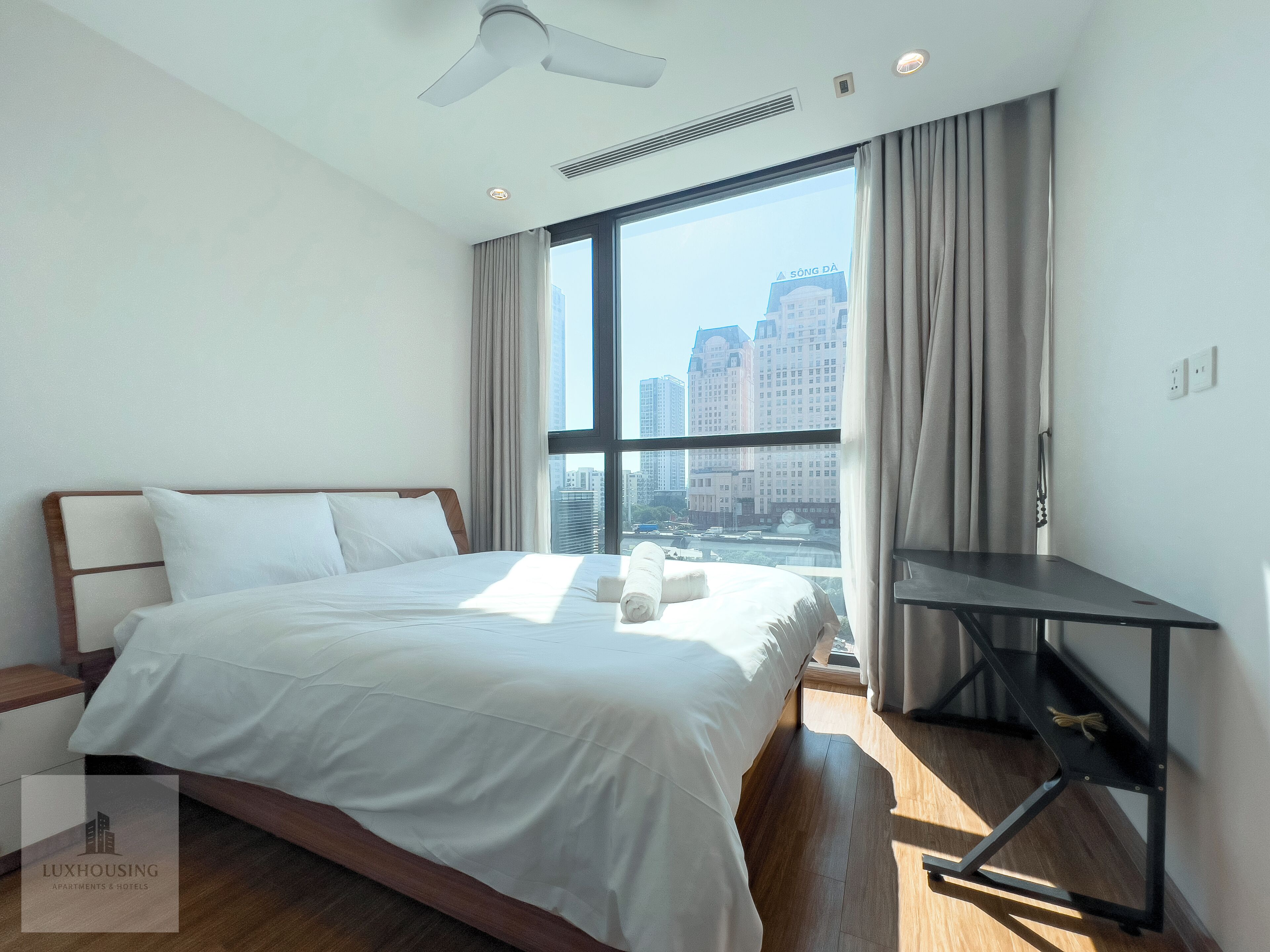 A modern hotel room with a large bed, a desk, and a view of the city skyline through a large window.