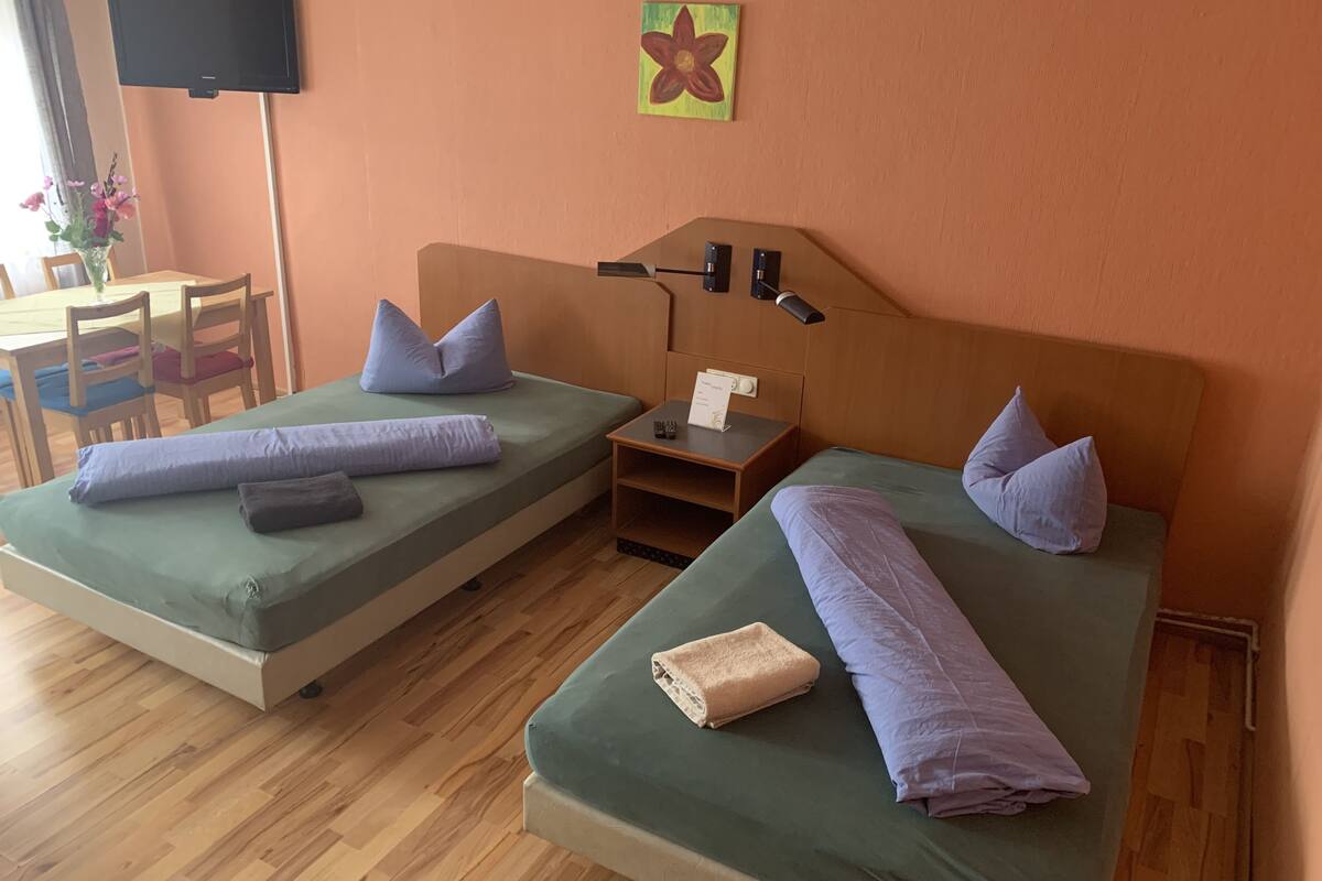 Two single beds with green mattresses, each with a pillow and a rolled towel. There is a small table with a sign, a wall-mounted TV, and a flower painting.
