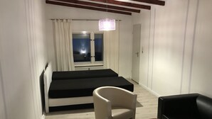 A compact living space with a sofa, a chair, a small table, and a window with curtains. - Pension Scheffler (Potsdam)
