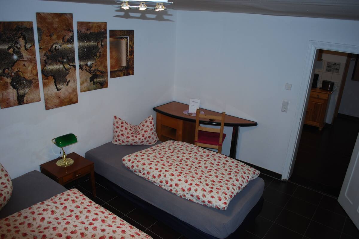 A small, cozy bedroom with a single bed, a desk, and a chair. There are three wall-mounted paintings and a green desk lamp.