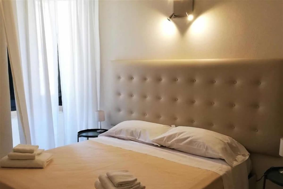 A neatly made bed with a tufted headboard, a bedside table with a lamp, and a window with sheer curtains.