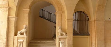 A grand staircase with classical architecture, flanked by two lion statues, leading to a balcony with a wrought iron railing.