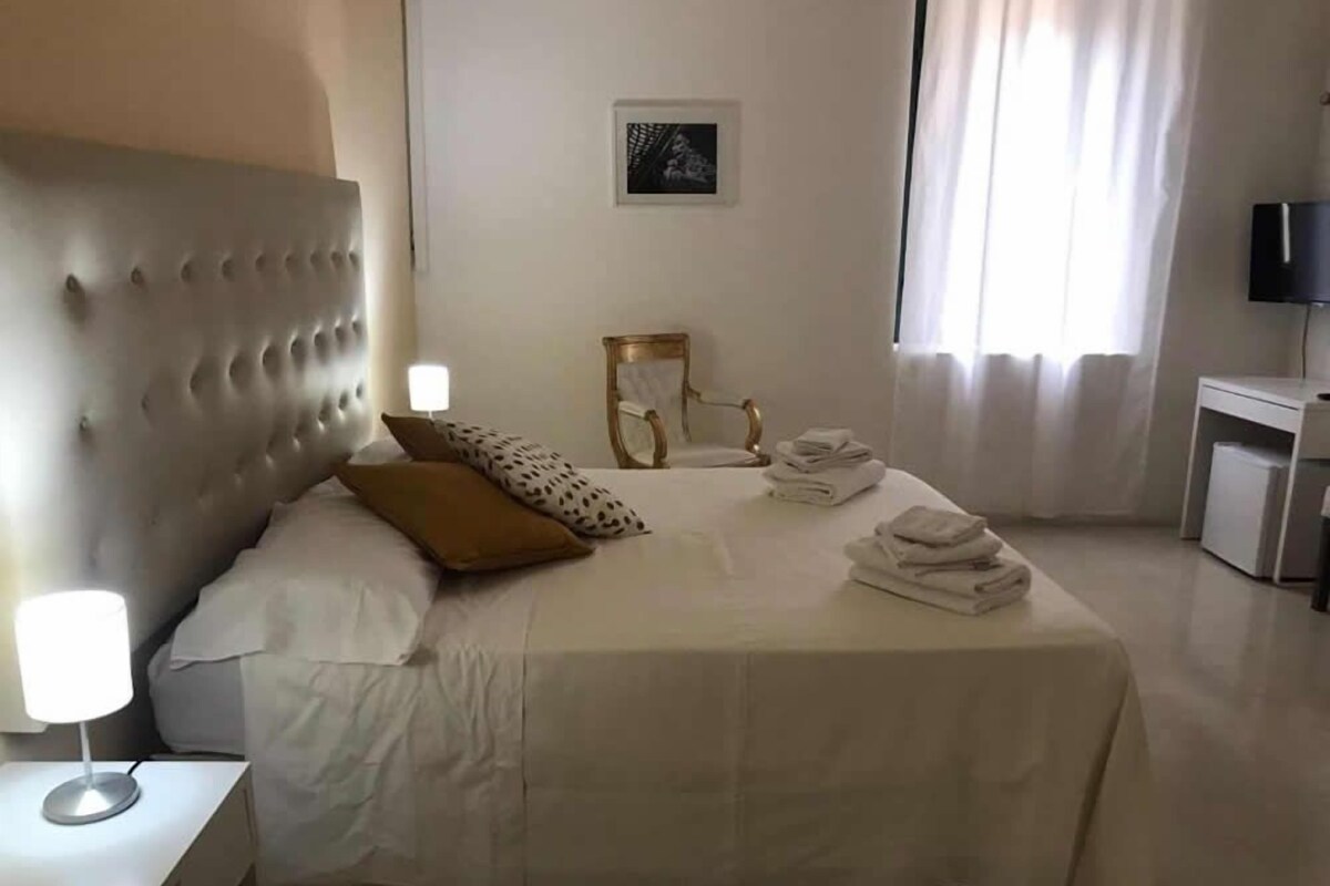 Una cama bien hecha con cabecera, una lámpara de noche y una silla. Hay una mesita con una lámpara, una toalla doblada y un cuadro en la pared.