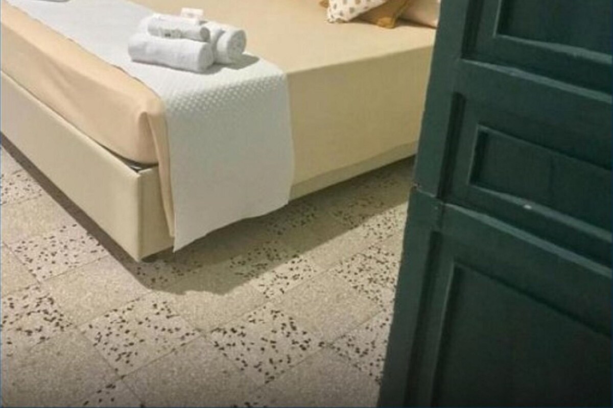 Una cama bien hecha con cabecera color beige, almohadas blancas y una manta blanca. Hay toallas sobre la cama y una pequeña mesa con una lámpara al lado.