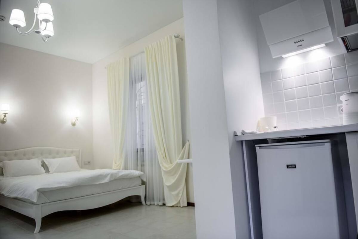 A bedroom with a bed, a window with sheer curtains, a wall-mounted air conditioner, and a small refrigerator.