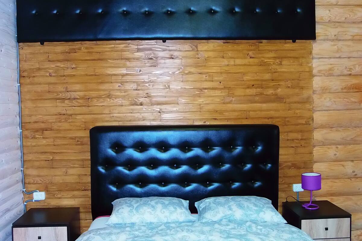 Een slaapkamer met een gestoffeerd hoofdeinde, houten wanden, een bed met een decoratief sprei en een paarse lamp op een nachtkastje.
