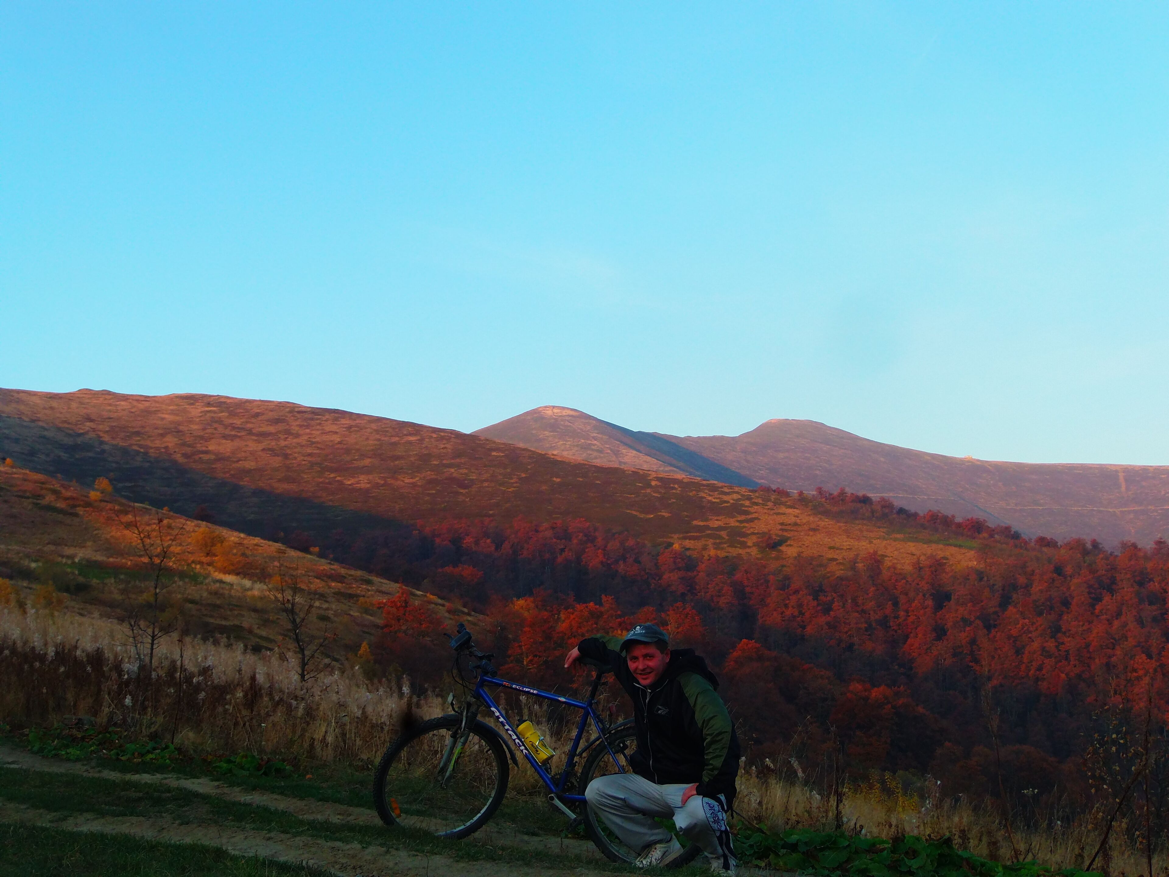 A person posing with a bicycle on a dirt path with a mountainous backdrop.