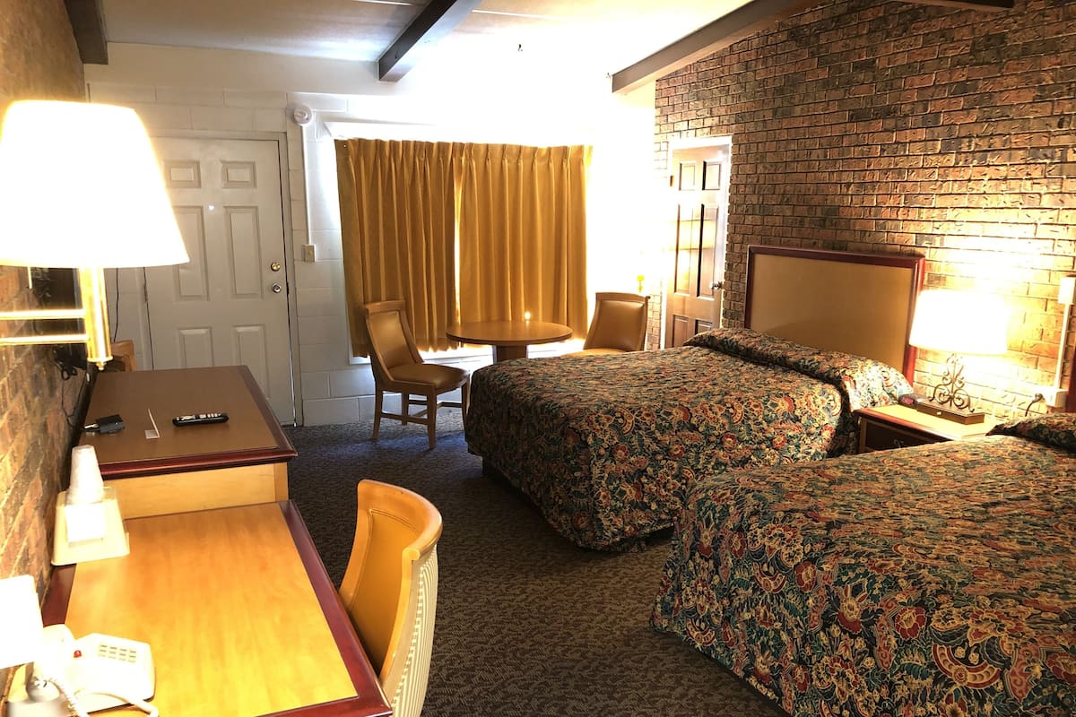 Quarto de hotel com escrivaninha de madeira, duas camas de solteiro com roupa de cama floral, uma cadeira, uma mesinha e uma parede de tijolos.