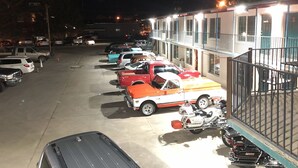 Un parking où sont stationnés plusieurs véhicules, dont des voitures et une moto, devant un immeuble doté de nombreuses fenêtres et d’un balcon.