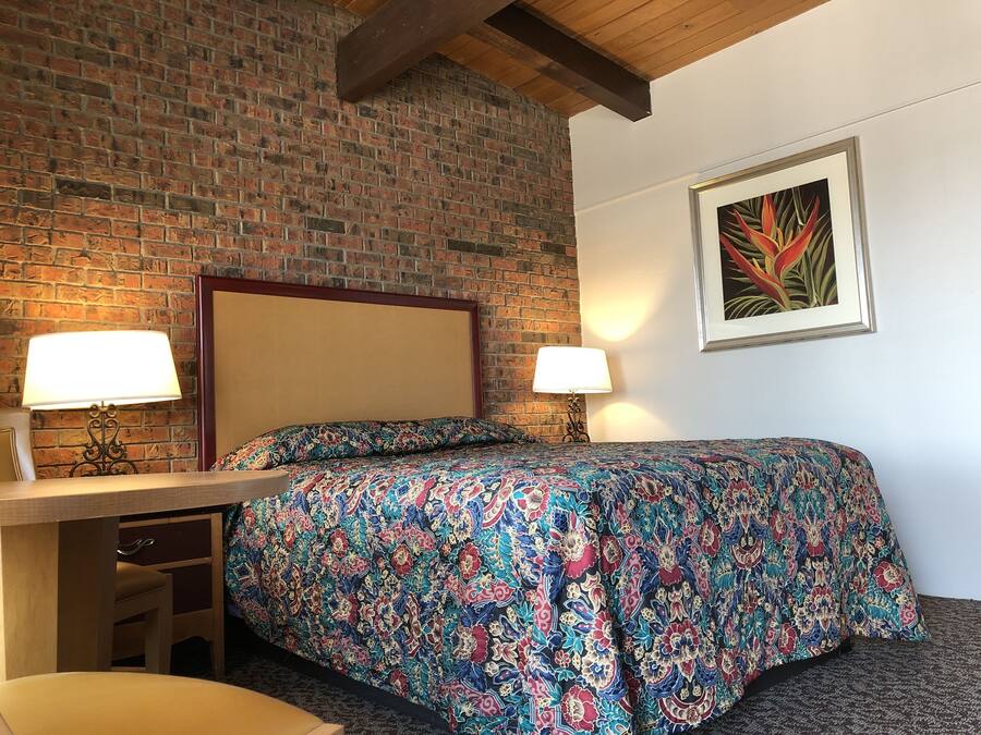 A hotel room with a brick wall, a bed with a colorful floral bedspread, two bedside lamps, a wooden desk with a chair, and a framed picture on the wall.