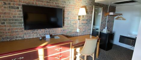 Una habitación con pared de ladrillo, un televisor de pantalla plana sobre un escritorio de madera con cajones, una silla y una lámpara.