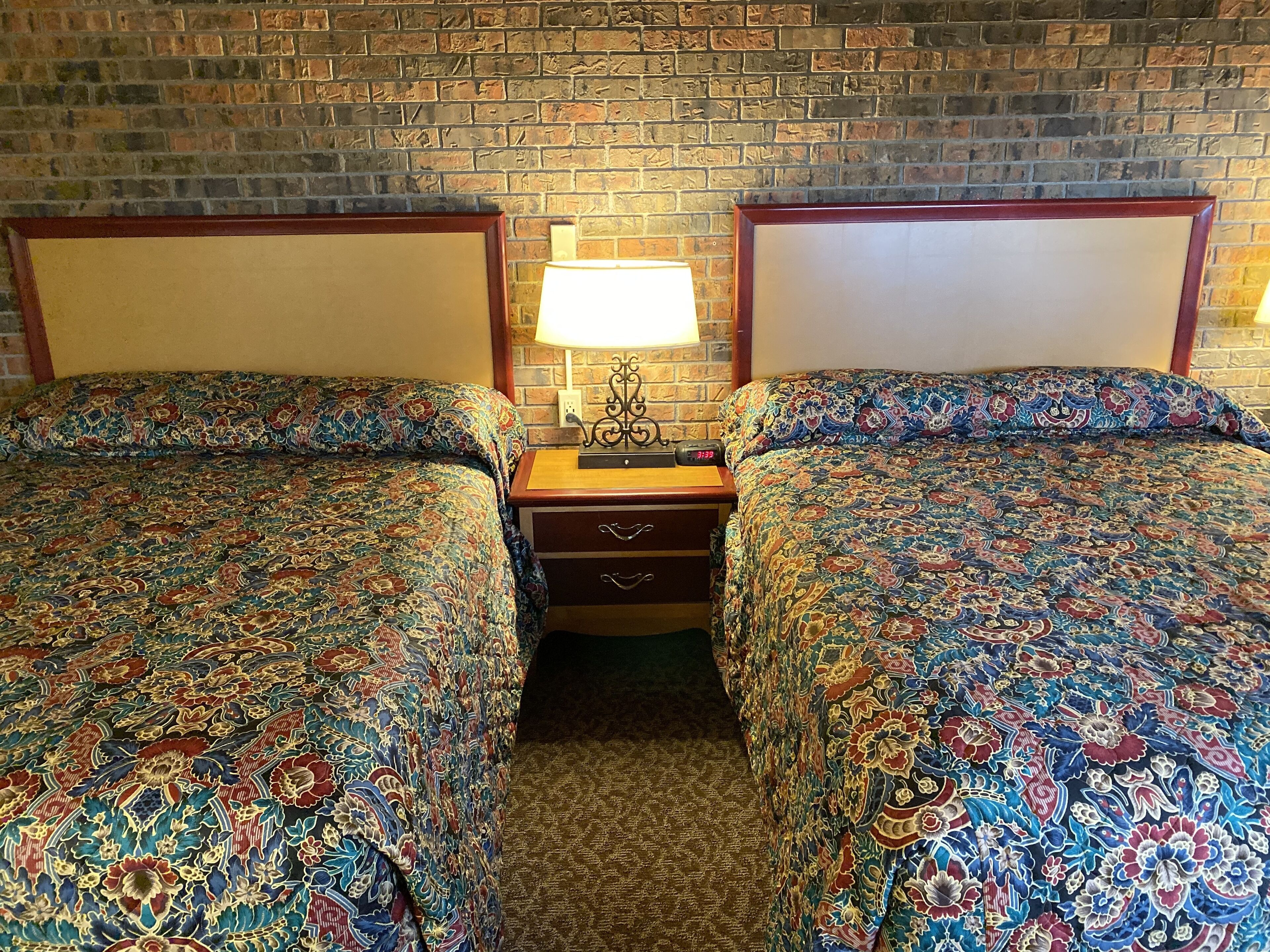 Ein Hotelzimmer mit zwei Betten, jedes mit einem gemusterten Überwurf, einem Nachttisch mit Lampe und einer Ziegelwand.