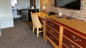 A hotel room with a brick wall, a flat-screen TV mounted on the wall, a wooden dresser with drawers, a desk with a chair, a lamp, and a kitchenette area with a refrigerator and sink.