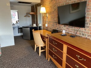 A hotel room with a brick wall, a flat-screen TV mounted on the wall, a wooden dresser with drawers, a desk with a chair, a lamp, and a kitchenette area with a refrigerator and sink.