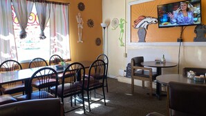L’intérieur d’un restaurant présente une salle à manger avec des tables et des chaises, un téléviseur fixé au mur et des œuvres d’art décoratives.