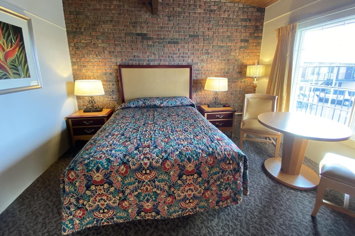 Quarto de hotel com parede em padrão tijolo, uma cama grande com colcha estampada, duas mesas de cabeceira com luminárias, uma mesa redonda com uma cadeira e uma janela com vista para a rua.