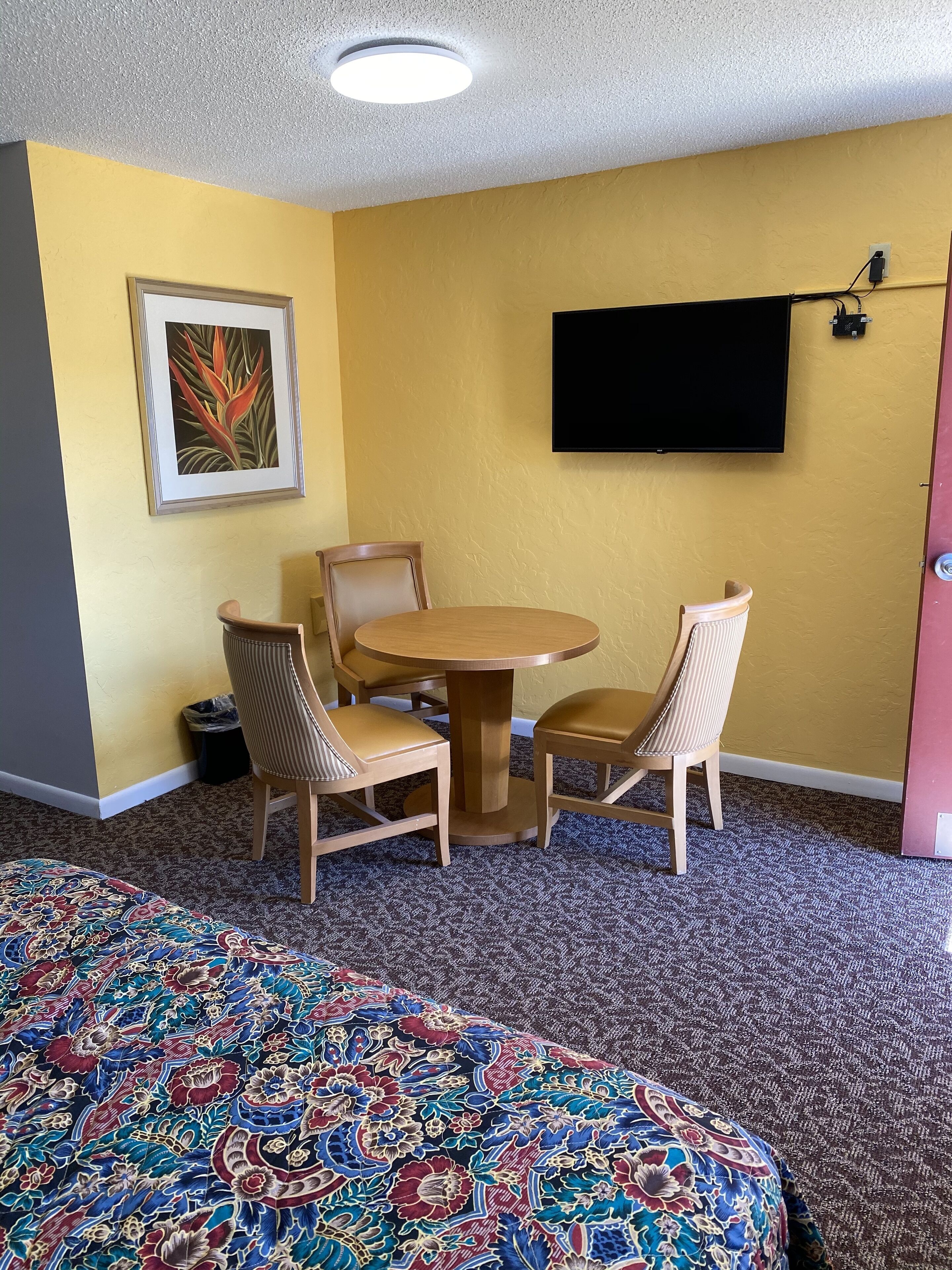 Une chambre d’hôtel avec un lit, une table ronde entourée de quatre chaises, un tableau encadré accroché au mur et un téléviseur à écran plat fixé au mur.